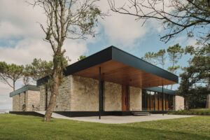 Hlavní vstup do domu zvýrazňuje kryté zápraží, kamenné stěny a elegantní dřevěné dveře pod širokým přesahem střechy.