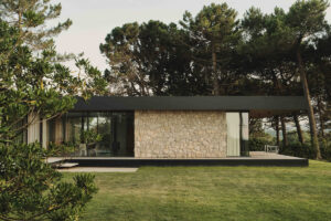 Pohled ze zahrady na dům ukazuje kombinaci kamenné fasády, velkého prosklení a terasy otevřené do zeleně.