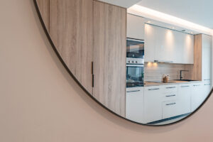 Kulaté zrcadlo v interiéru odráží moderní kuchyňskou linku se sporákem a pracovní deskou v přírodních tónech.