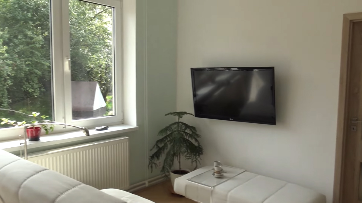 Minimalistická obývací část s nástěnnou televizí a jednoduchým nábytkem využívá přirozené světlo z velkého okna.