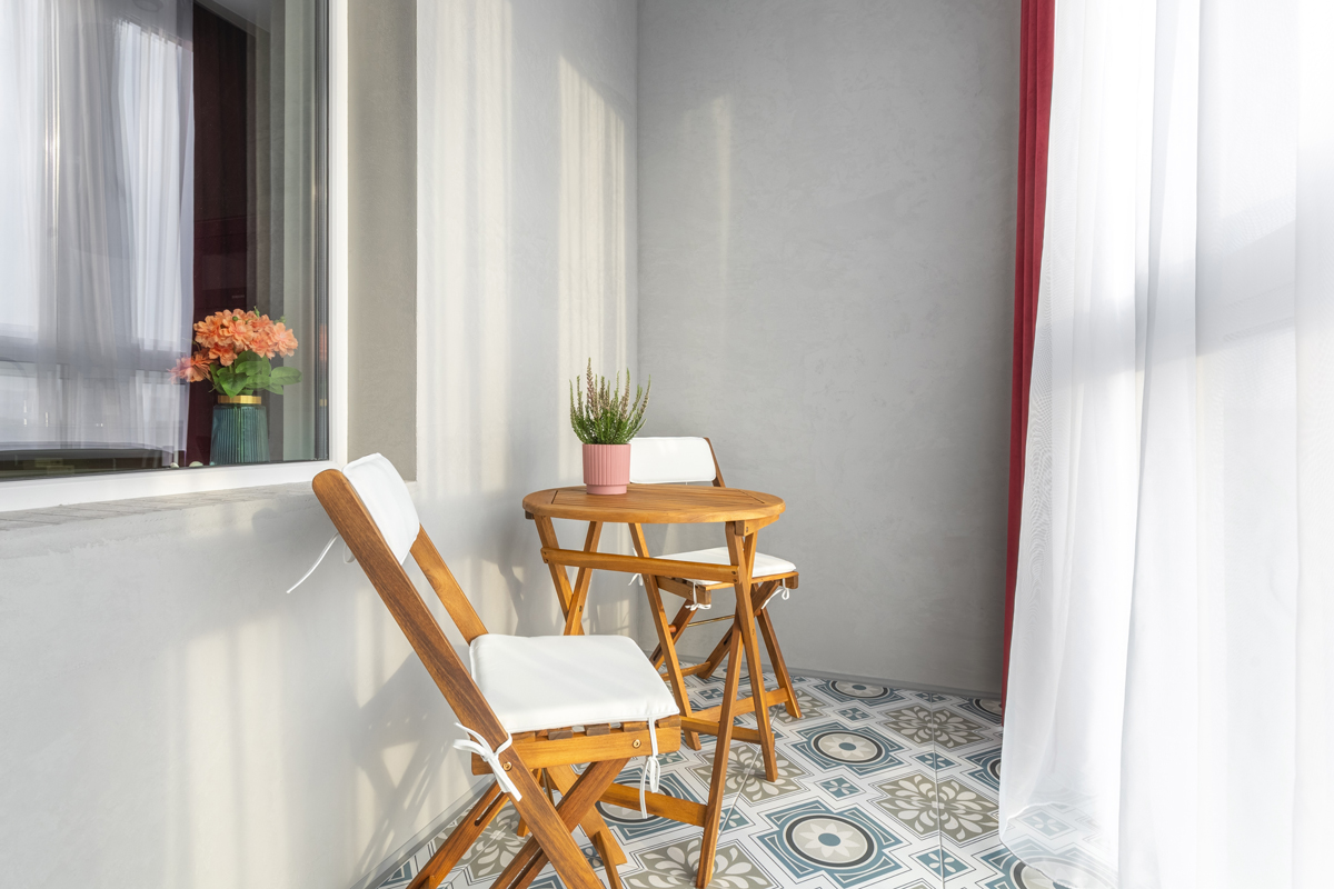 Úzký balkon s dřevěným skládacím nábytkem, světlými textiliemi a dekorativní dlažbou ukazuje, jak mohou přírodní materiály sjednotit venkovní prostor s interiérem bytu.