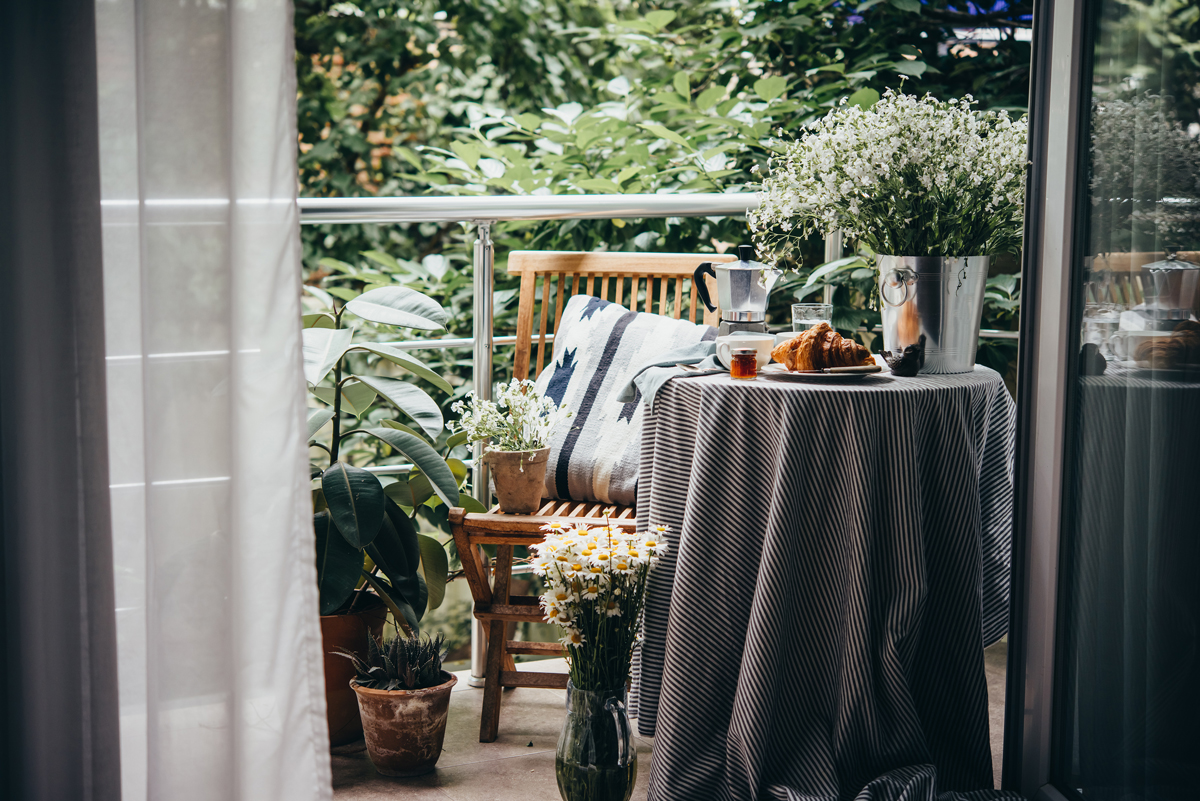Útulný balkon s malým stolkem, croissantem, květinami a množstvím zeleně připomíná venkovní obývací pokoj a ukazuje, jak důležitou roli hrají textilie, rostliny a dekorace při propojení bytu s exteriérem.
