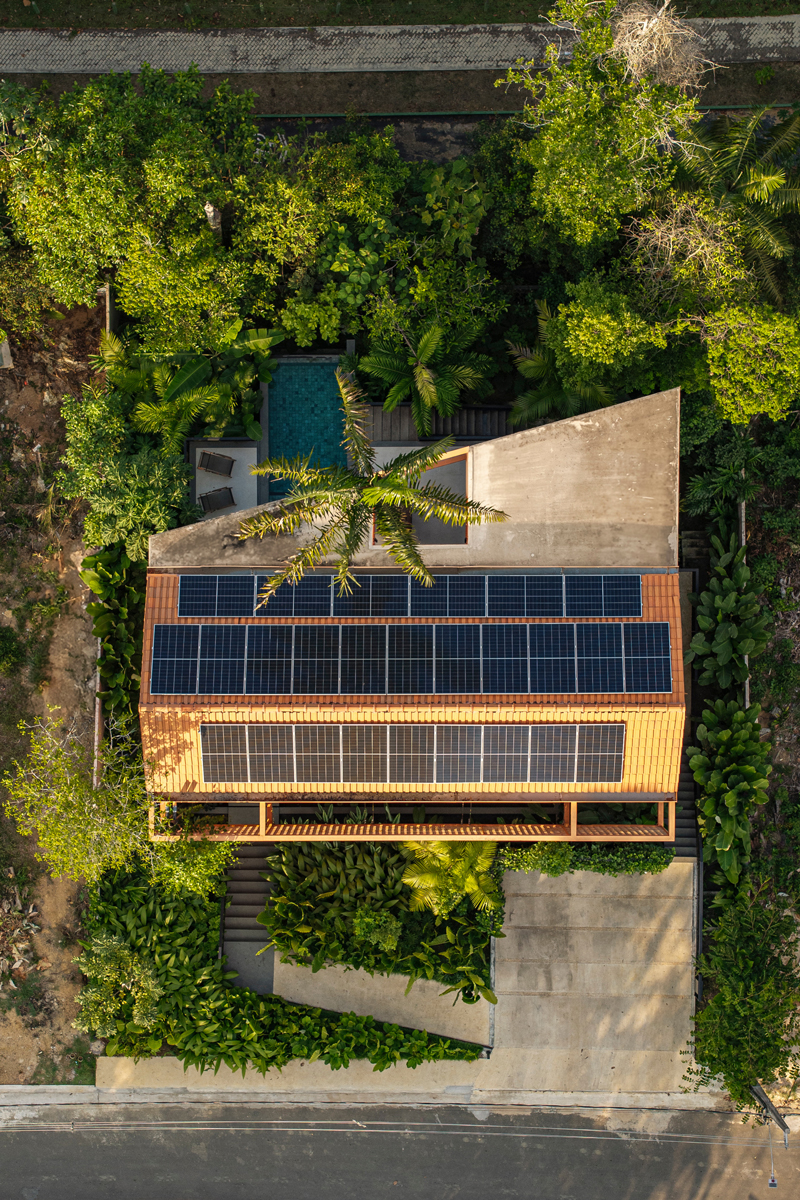 Pohled z výšky ukazuje střechu domu se solárními panely obklopenou hustou tropickou vegetací.
