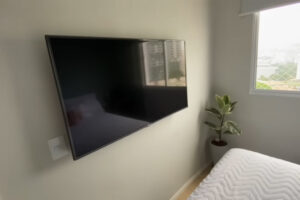 Ložnice využívá minimalistické zařízení s televizí na stěně a malým pokojovým stromkem u okna.