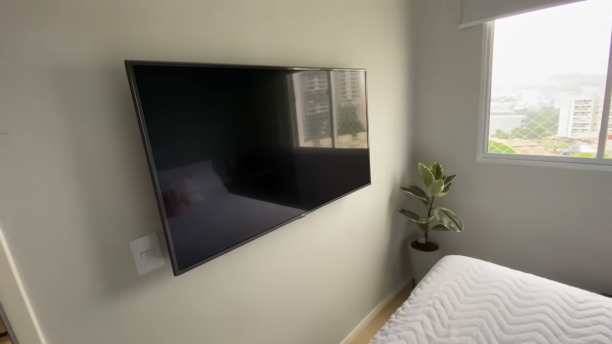Ložnice využívá minimalistické zařízení s televizí na stěně a malým pokojovým stromkem u okna.