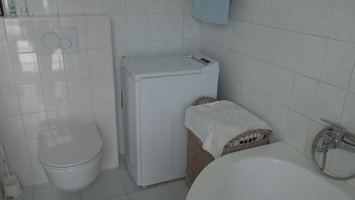 Koupelna kombinuje závěsnou toaletu, pračku a vanu v jednom prostoru s původními bílými obklady.
