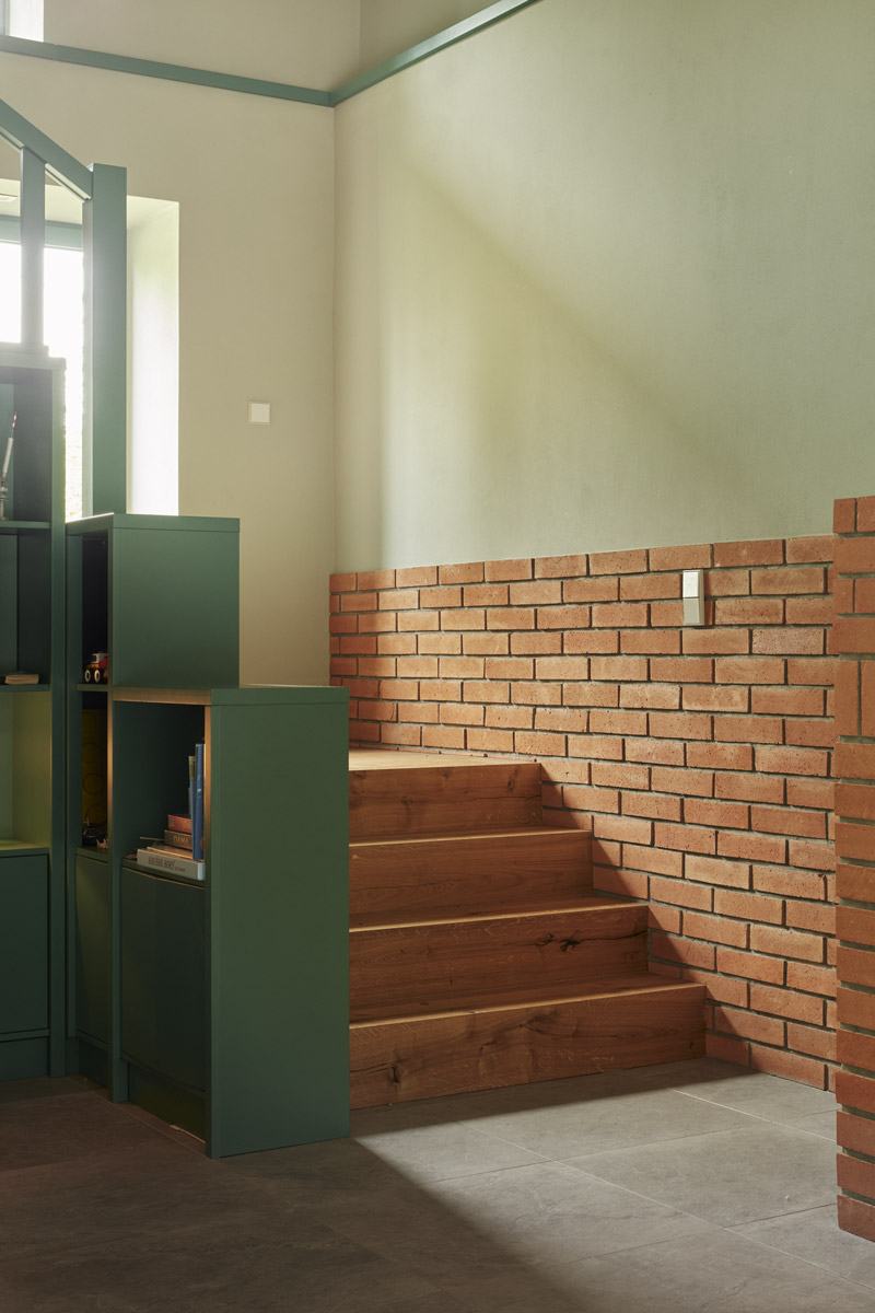 Dřevěné schodiště navazuje na zelenou knihovnu a cihlovou stěnu, které společně určují charakter hlavního obytného prostoru.