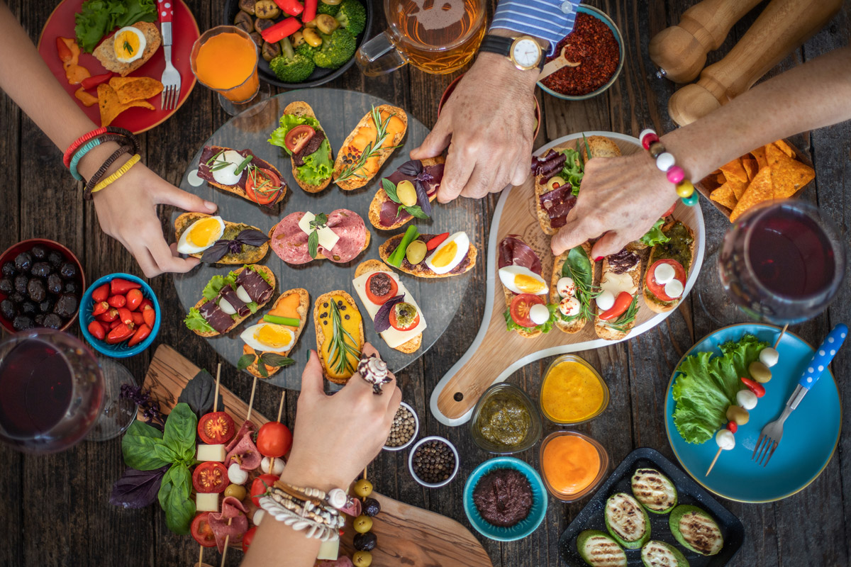 Pestrý výběr tapas, drobných obložených chlebíčků, oliv, zeleniny a omáček rozložený na stole při společném posezení.