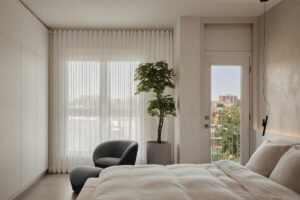 Světlá ložnice s velkým oknem, pohodlným křeslem a přístupem na balkon působí vzdušně a klidně.