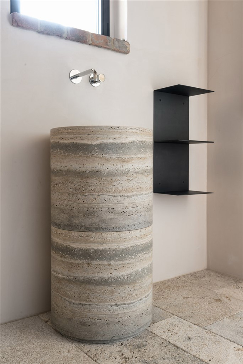 Vysoké válcové umyvadlo z vrstveného travertinu s nástěnnou baterií a černou kovovou policí.