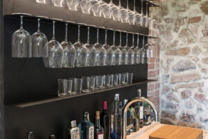 Domácí bar s černou kovovou konstrukcí na sklenice, kamennou pracovní deskou a malým dřezem.