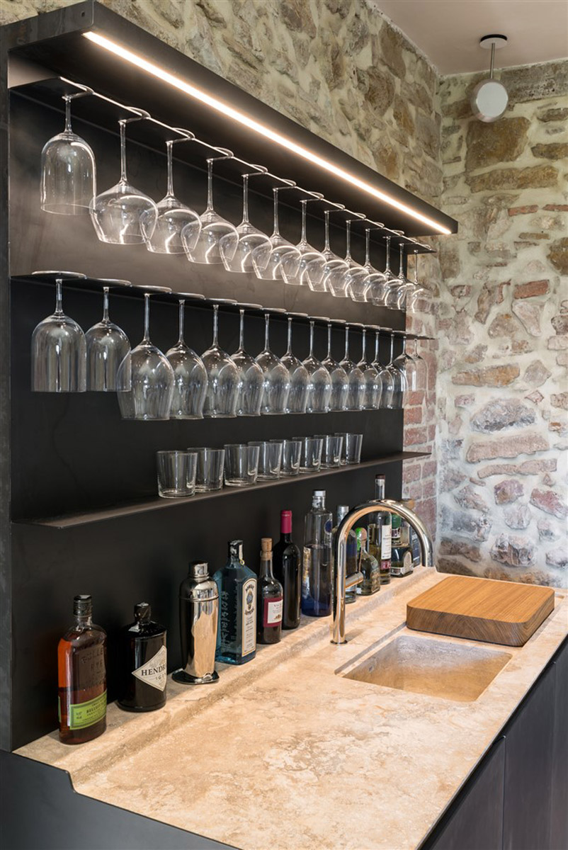 Domácí bar s černou kovovou konstrukcí na sklenice, kamennou pracovní deskou a malým dřezem.