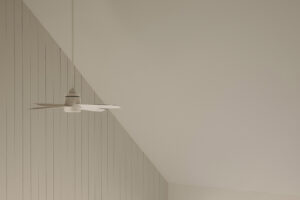 Vysoký světlý interiér se šikmým stropem, závěsným ventilátorem a velkými okny do krajiny.