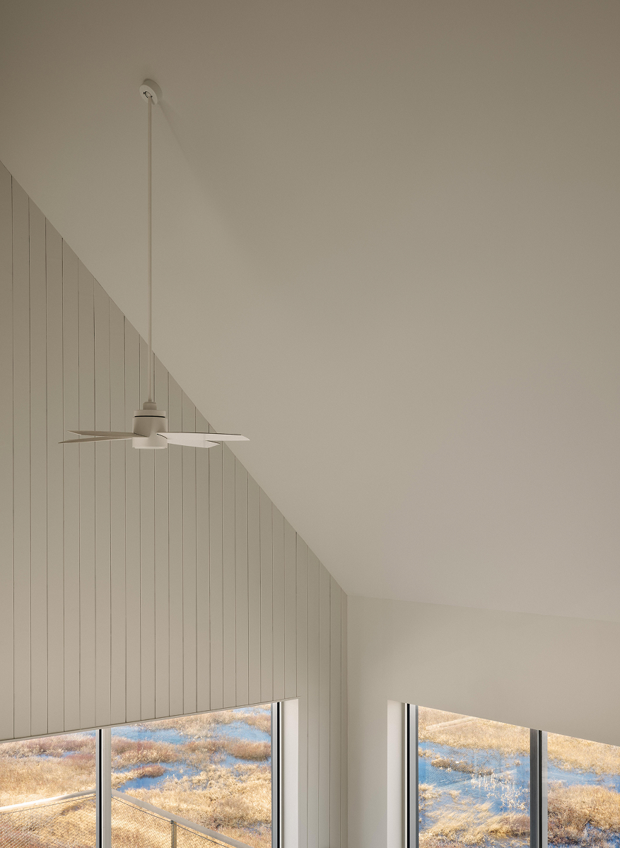 Vysoký světlý interiér se šikmým stropem, závěsným ventilátorem a velkými okny do krajiny.