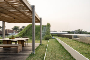 Dřevěná pergola s venkovním jídelním stolem na zelené střeše vedle travnatého svahu se schodištěm.