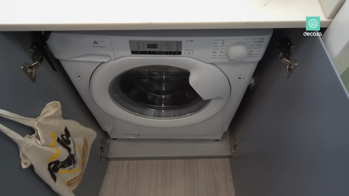 Pračka ukrytá za modrošedými dvířky kuchyňské linky v bytě o rozloze 16 metrů čtverečních.