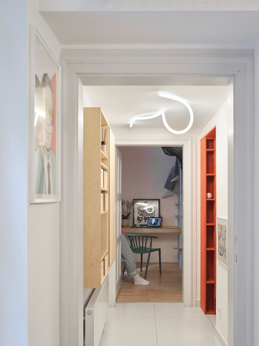 Úzká světlá chodba s vestavěným nábytkem ze dřeva, červenými policemi a pracovním koutem v pozadí.