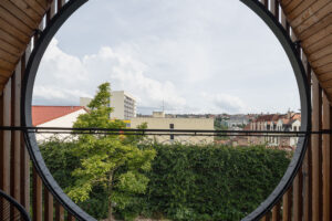 Dřevěný balkon s velkým kruhovým otvorem a výhledem na okolní zástavbu a zeleň.