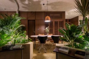 Pracovní kout a jídelnu lemují vestavěné květináče s tropickou zelení, zatímco tmavé dřevo nábytku sjednocuje otevřený interiér.