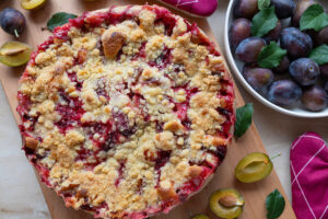 Upečený švestkový crumble se zlatavou drobenkou obklopený čerstvými a rozpůlenými švestkami.