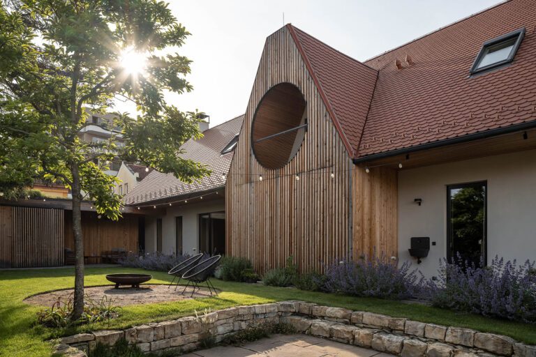 Zahrada u domu s dřevěnou fasádou, kruhovým otvorem, trávníkem, posezením a kamennými zídkami.
