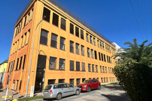 Pohled na čtyřpatrovou budovu DADA Distriktu v Brně se zlatavě žlutou fasádou, pravidelnými řadami oken a zaparkovanými auty před vstupem.