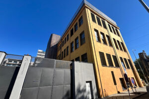 Rohový pohled na zrekonstruovanou industriální budovu DADA Distriktu se žlutou omítkou, vysokými okny a oploceným prostorem u vstupu.