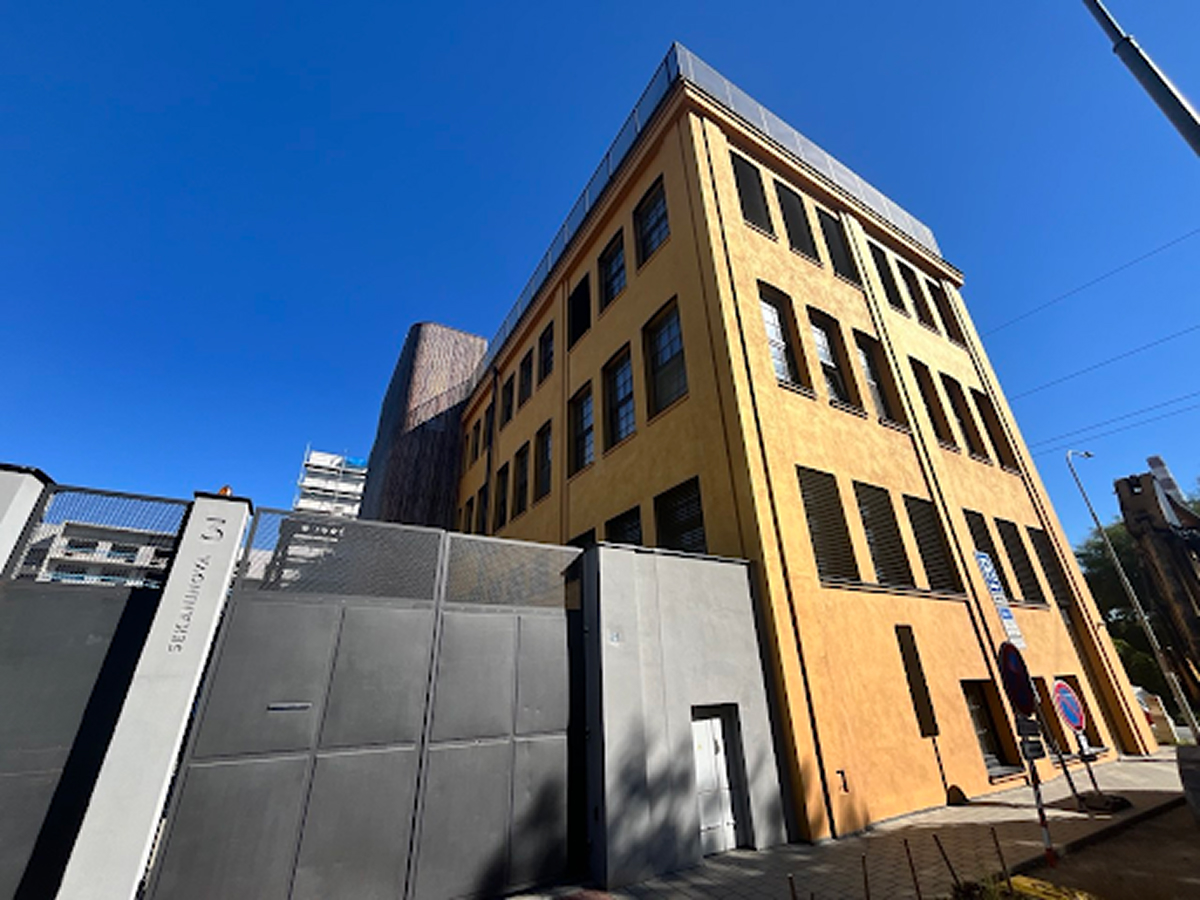Rohový pohled na zrekonstruovanou industriální budovu DADA Distriktu se žlutou omítkou, vysokými okny a oploceným prostorem u vstupu.