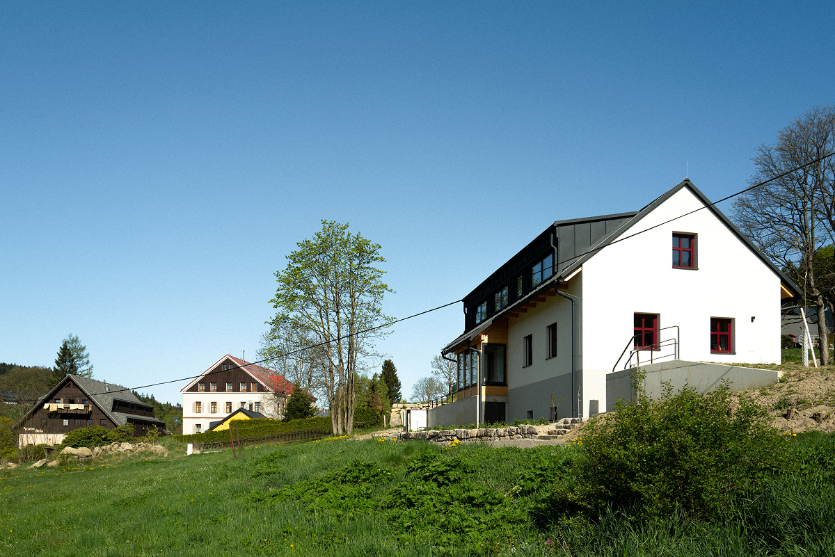 Boční pohled na horský rodinný dům v Bedřichově s bílou štítovou fasádou, červenohnědými rámy oken, tmavou střechou a prosklenou verandou.