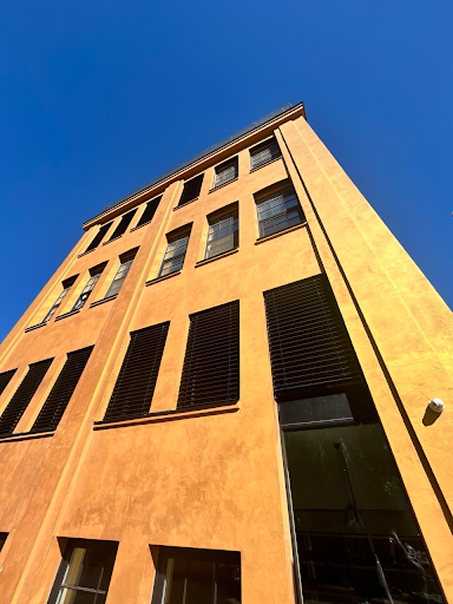 Detail vysoké žluté fasády DADA Distriktu v Brně s černými okenními rámy a venkovními žaluziemi, fotografovaný z podhledu proti modré obloze.
