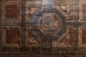 Detail historického dřevěného stropu s malovanými geometrickými ornamenty a figurální výzdobou v hnědých a tlumených zemitých odstínech.