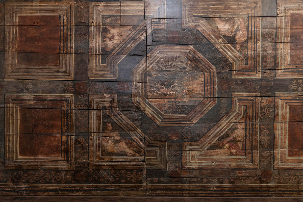 Detail historického dřevěného stropu s malovanými geometrickými ornamenty a figurální výzdobou v hnědých a tlumených zemitých odstínech.