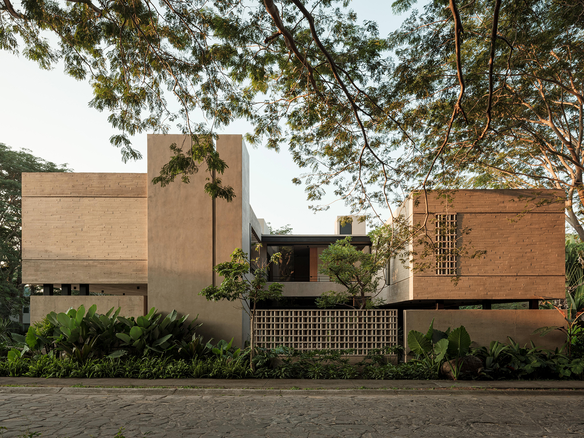 Pohled z ulice na moderní betonový dům Casa Akina s několika hranolovými částmi, perforovanými stěnami a bujnou zelení.