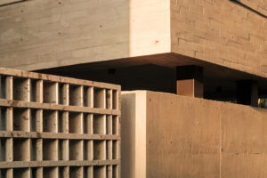 Detail rohu moderního domu z pohledového betonu s geometrickými mřížkami, betonovou ohradní zdí a zelenými rostlinami.
