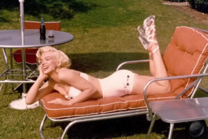 Marilyn Monroe v bílých plavkách a sandálech odpočívá na oranžovém zahradním lehátku vedle kovového stolku na slunném trávníku.