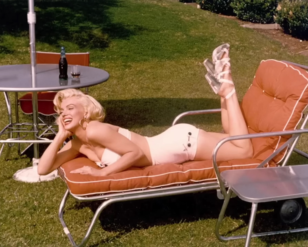 Marilyn Monroe v bílých plavkách a sandálech odpočívá na oranžovém zahradním lehátku vedle kovového stolku na slunném trávníku.