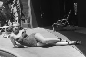 Černobílá fotografie Marilyn Monroe v tmavých plavkách a sandálech, jak pózuje vleže na terase vedle bazénu.