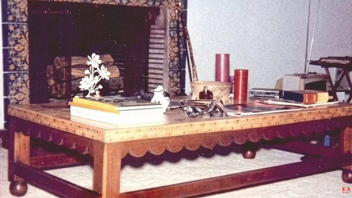 Historický snímek interiéru s nízkým dřevěným konferenčním stolkem, knihami a krbem obloženým dekorativními keramickými dlaždicemi.