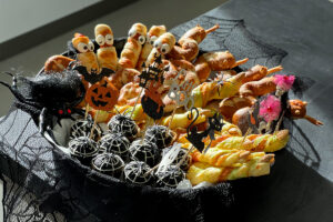 Talíř s halloweenským občerstvením pro děti, na kterém jsou sladkosti zdobené jako mumie a pavučiny, slané pečivo a strašidelné dekorace.