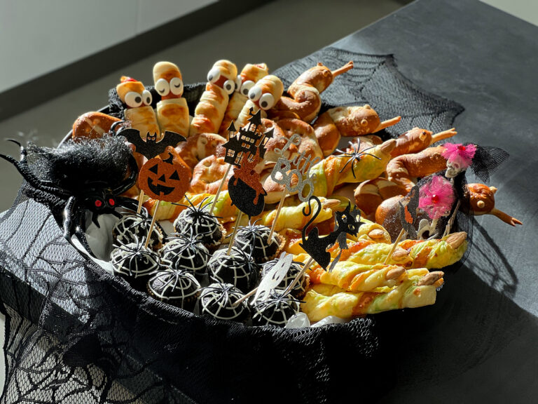 Talíř s halloweenským občerstvením pro děti, na kterém jsou sladkosti zdobené jako mumie a pavučiny, slané pečivo a strašidelné dekorace.