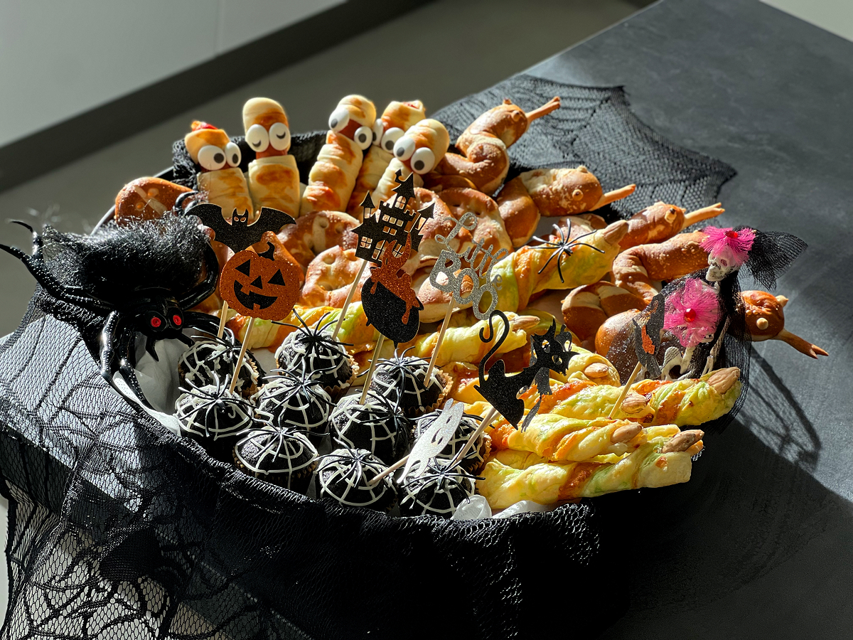 Talíř s halloweenským občerstvením pro děti, na kterém jsou sladkosti zdobené jako mumie a pavučiny, slané pečivo a strašidelné dekorace.