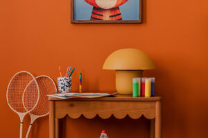 Dětský kreativní koutek s oranžovou stěnou, dřevěným stolkem, výtvarnými potřebami, stolní lampou a obrázkem tygra.