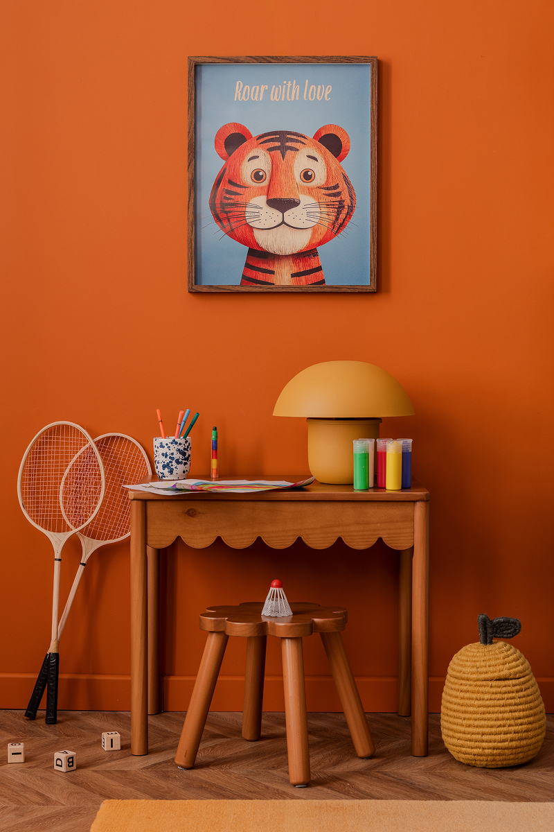 Dětský kreativní koutek s oranžovou stěnou, dřevěným stolkem, výtvarnými potřebami, stolní lampou a obrázkem tygra.