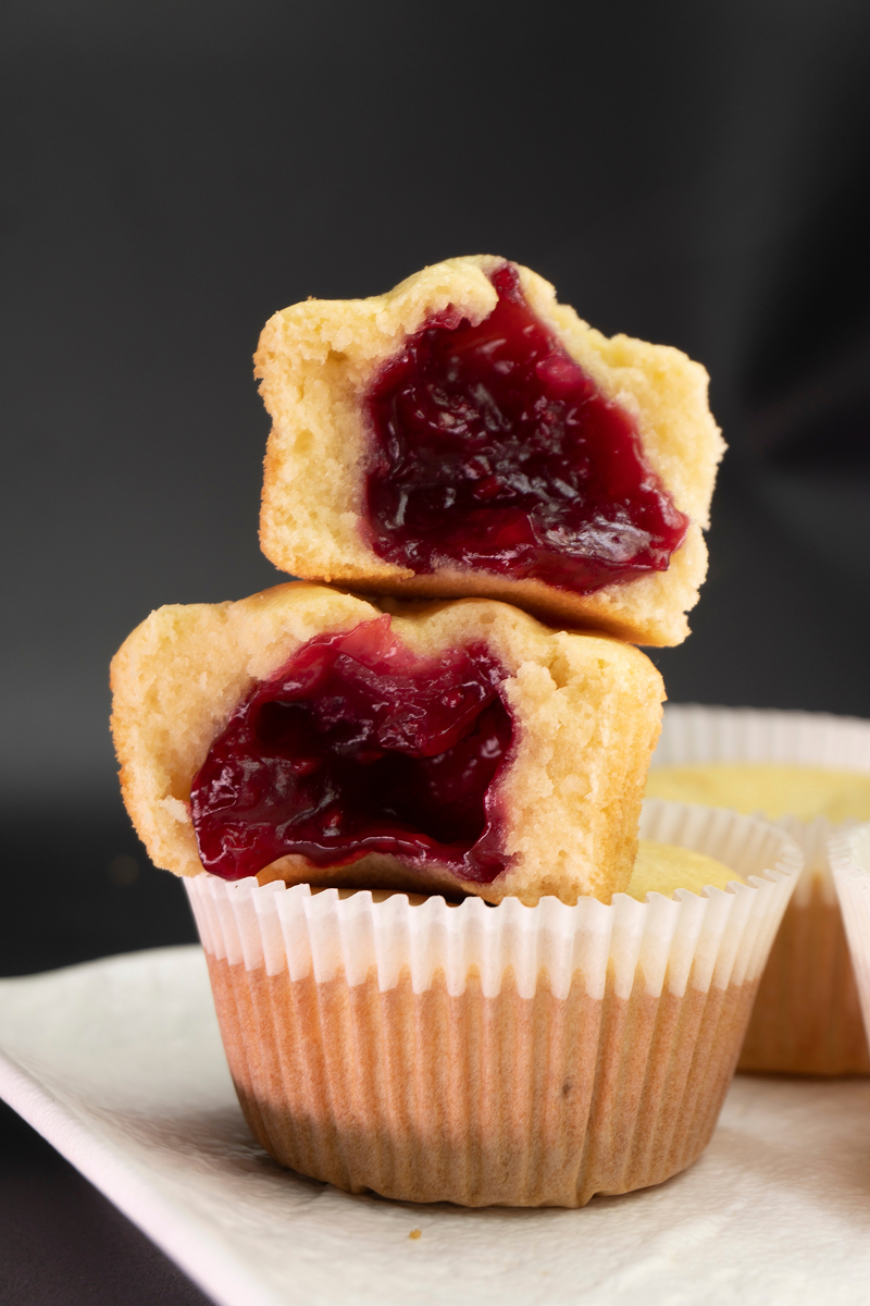 Rozkrojené muffiny se světlým těstem a výraznou červenou ovocnou náplní, která vytéká ze středu.