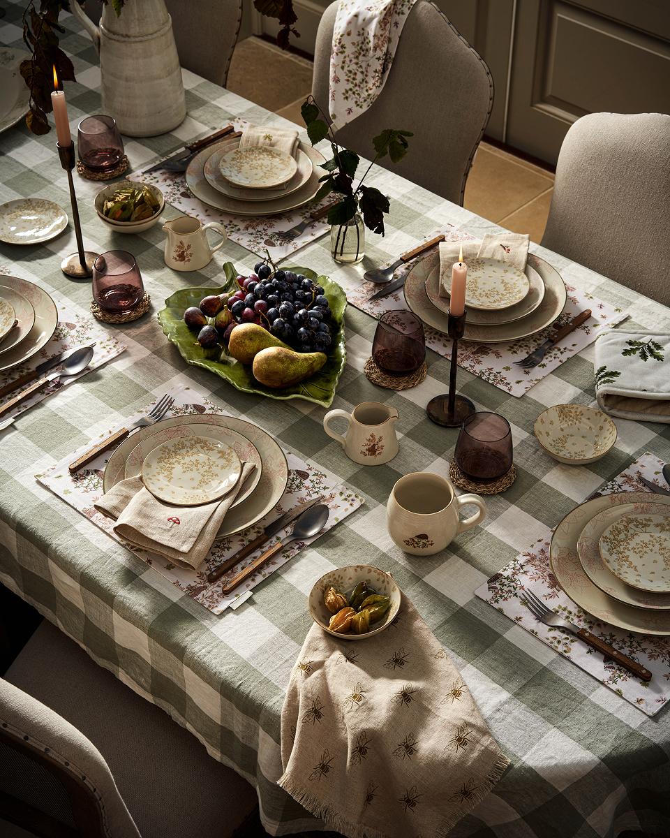 Podzimní jídelní stůl se zeleným kostkovaným ubrusem, květovaným porcelánem, fialovými skleničkami, svíčkami a mísou hrušek a hroznového vína.